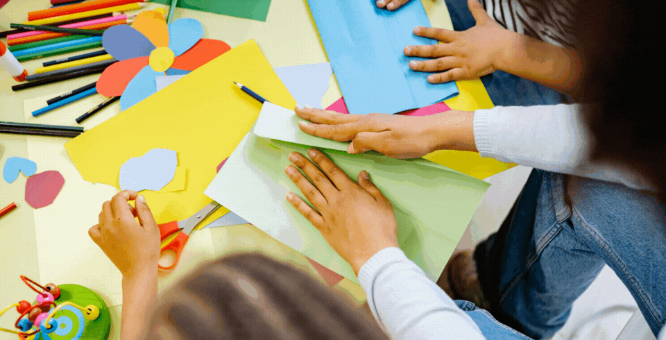 two children and one adult fold brightly coloured paper on a table